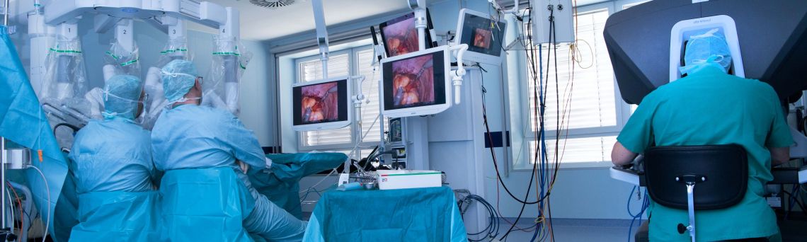 Blick in einen OP-Saal, in dem eine Roboter-assistierte Operatio durchgeführt wird. Drei Operateure sitzen mit dem Rücken zur Kamera. 
