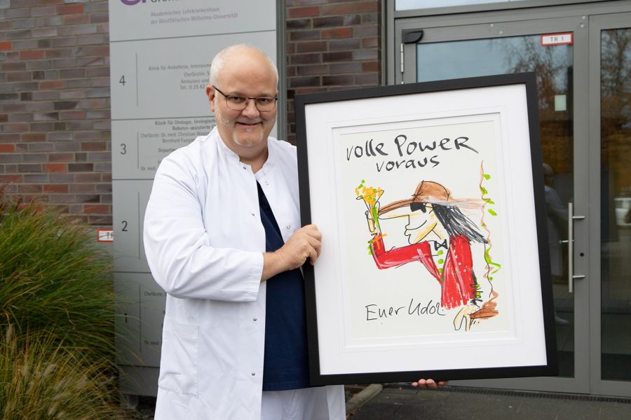 Chefarzt Dr. Christian Wagner steht mit dem Gemälde von Udo Lindenberg im Arm vor dem Eingang des Krankenhauses und lächelt in die Kamera.  