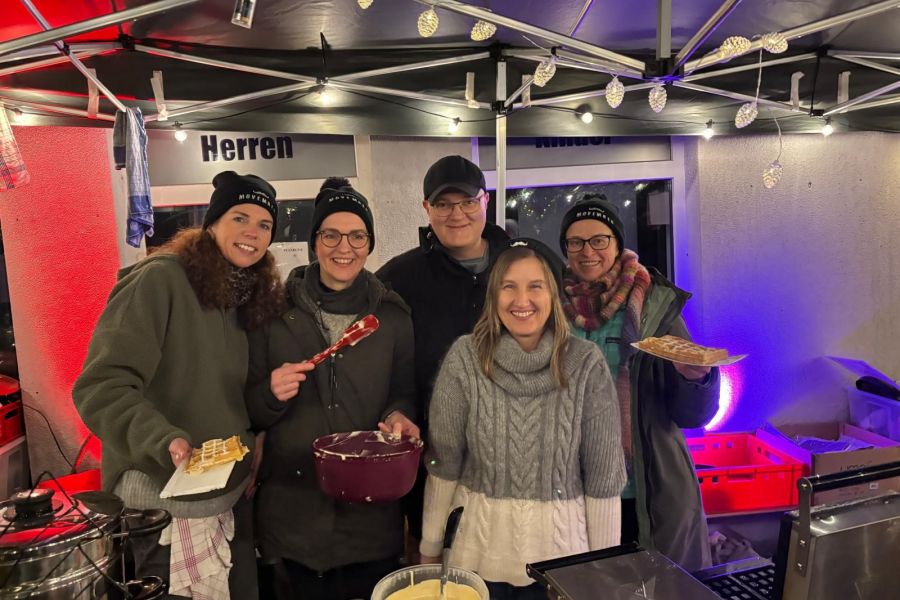 Part of the “Beards Without Borders” team – Dr. Christian Wagner and four women – are standing behind the counter of the Christmas market booth in Bad Bentheim, smiling at the camera while selling waffles.