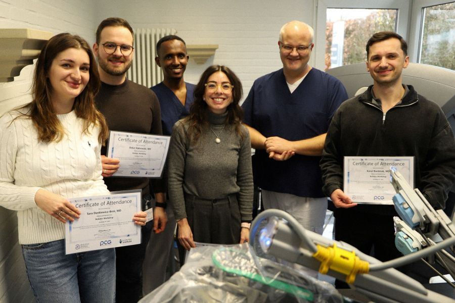 The four participants of the course with their certificates, along with Chief Physician Dr Christian Wagner (2nd from right) and Senior Physician Abdirahman Ayanle (3rd from left) behind the arms of a da vinci 5 Trainingssystem, smiling.   